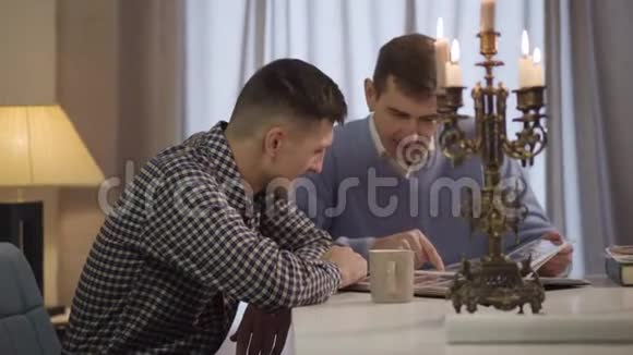 侧面观看成年英俊的白种人男子坐在桌子旁与中年父亲交谈儿子和父母视频的预览图