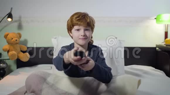 前景肖像可爱的白种人红头男孩坐在床上和切换频道与遥控器魅力视频的预览图
