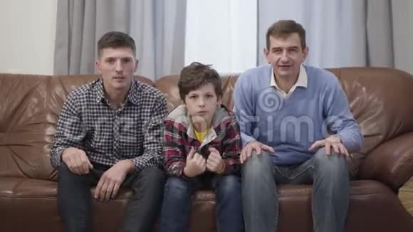 三个不同年龄的男人在电视上看足球比赛和打手势的肖像可悲的白人儿子父亲和视频的预览图