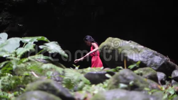 穿着红色衣服的年轻女子独自走在丛林中一个黑暗的洞穴附近视频的预览图