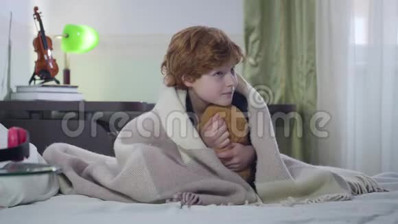 小红发白种人男孩抱着泰迪熊坐在床上晚上用毯子覆盖孩子童年视频的预览图