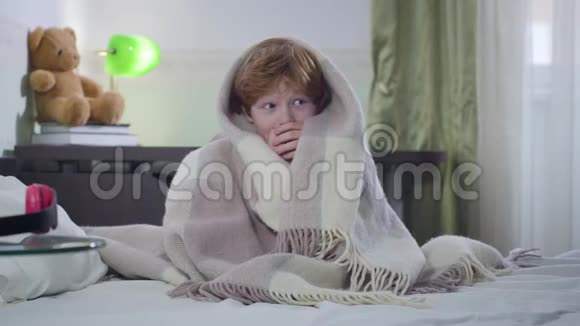 可爱的小白种人男孩坐在铺着毯子的床上环顾四周的肖像带着红色头发的孩子视频的预览图