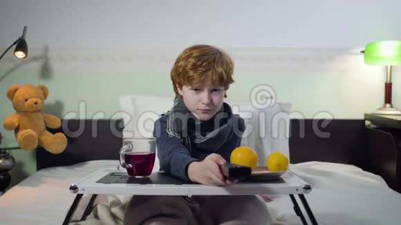 病态白人男孩用遥控器切换频道和看电视的肖像可爱的红头孩子坐在床上视频的预览图