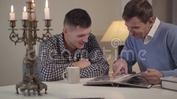 中年白种人的父亲和成年儿子坐在桌子旁看着相册里的照片友好的父母和男人视频的预览图