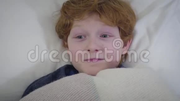 快乐的白种人红头孩子躺在床上躲在毯子下的肖像快乐的男孩去睡觉视频的预览图