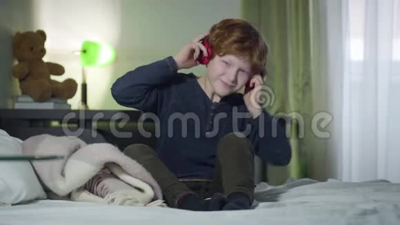 热情的白种人红发男孩在耳机听音乐快乐的小家伙在床上摇头视频的预览图