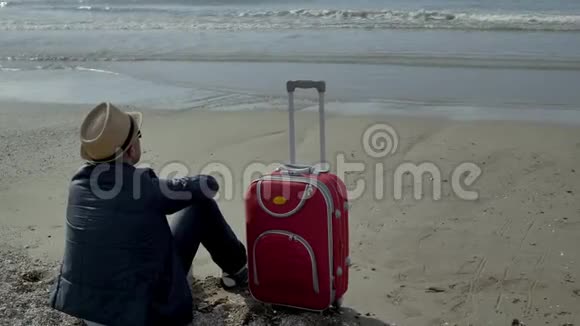 旅行者手里拿着一个红色的手提箱坐在海边的沙滩上休息积极的情绪视频的预览图