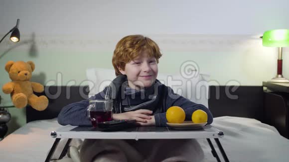 开朗的白人红发男孩看电视笑带遥控托盘的可爱生病的孩子坐在床上视频的预览图