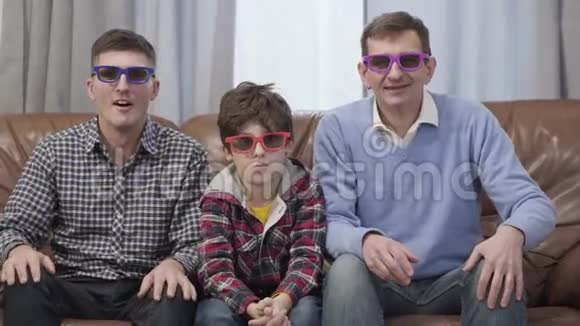 三个不同年龄的白人男子坐在家里的沙发上戴着3D护目镜看电视快乐的多代人视频的预览图