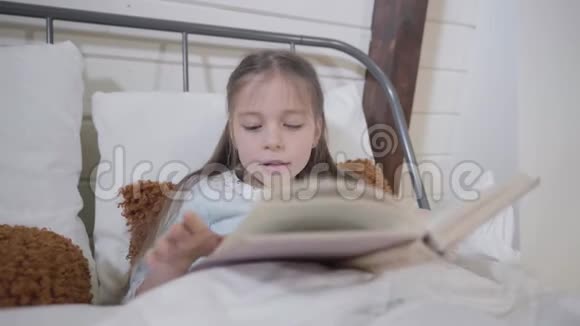 一幅美丽的白种人女孩躺在床上看书的肖像聪明的孩子晚上睡觉卧床时间视频的预览图