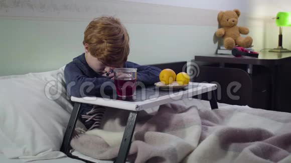 恶心的白种人男孩坐在托盘前与热饮料和橘子可怜可爱的红头孩子的肖像视频的预览图