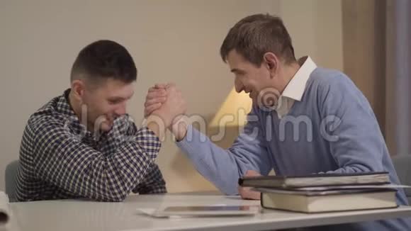 中年和成年白种人坐在桌子旁做手臂摔跤快乐微笑的儿子和父亲视频的预览图