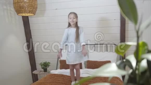一幅快乐的小白种人女孩在家玩得开心的肖像欢乐的孩子在室内的床上跳舞和跳跃视频的预览图