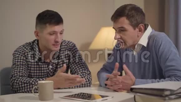 成年白种人儿子和中年父亲晚上在家谈话的肖像两个人坐在桌边视频的预览图