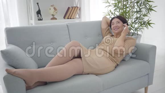优雅的中年白种人女人躺在沙发上在电话里聊天穿着米色连衣裙的漂亮老太太视频的预览图