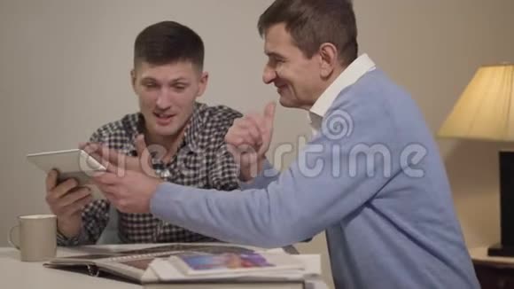侧面观看微笑的白种人中年男子使用平板电脑并与成年儿子在家交谈快乐的男人休息视频的预览图