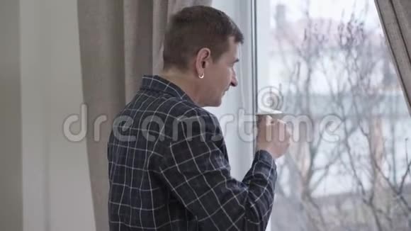 早上在窗前喝咖啡的带耳环的白种人成年男子的背面视图中年男子视频的预览图