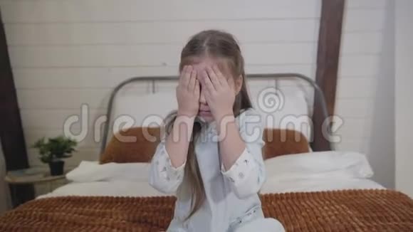 镜头靠近坐在床上的漂亮的白人小女孩她用手闭着眼睛开朗的孩子视频的预览图