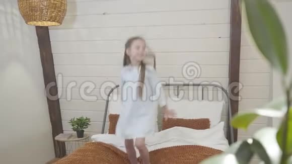 中镜头快乐的小白种人女孩跳在床上微笑快乐的黑发孩子在室内玩得开心视频的预览图