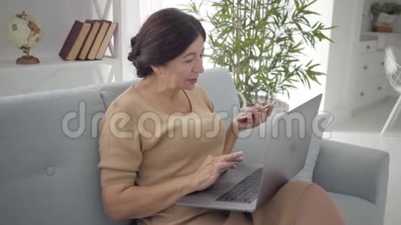 自信的高加索族妇女使用笔记本电脑和智能手机坐在室内沙发上成功的黑发中视频的预览图