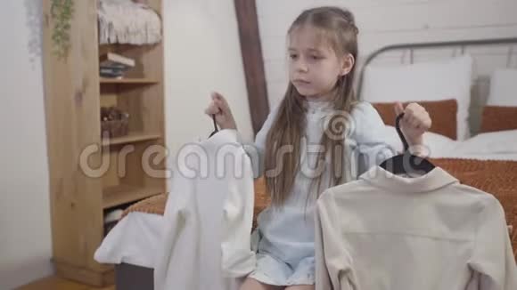 小白种人女孩坐在床上两个衣架和思考的肖像很漂亮的孩子会选择服装视频的预览图