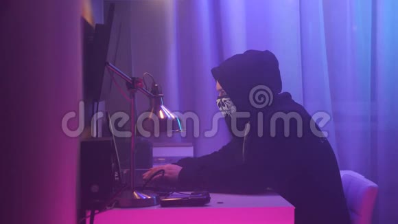 男性黑客坐在他的办公桌前闯入系统穿着黑色连帽衫戴着头巾的人把脸藏起来视频的预览图