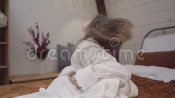 镜头接近可爱的小白种人女孩穿着白色浴袍摇着长长的卷发坐在床上的漂亮孩子视频的预览图