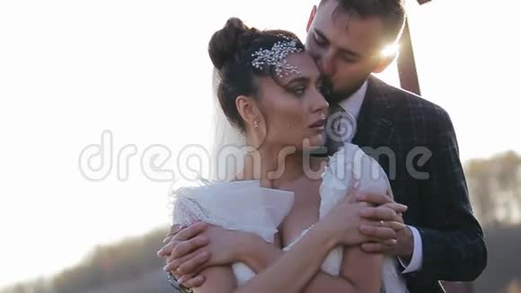 大胡子的新郎从背后拥抱亲吻他心爱的新娘在一片荒芜的田野里阳光明媚视频的预览图