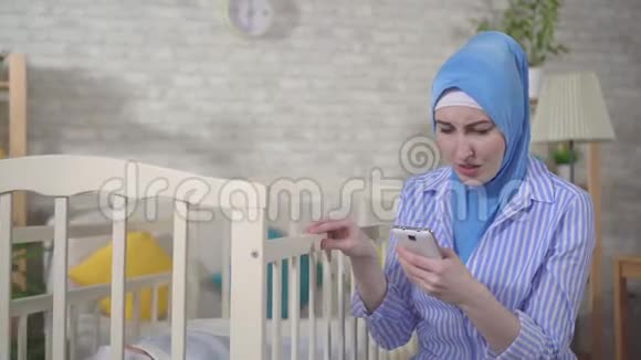 焦虑的穆斯林年轻女子是一位母亲在婴儿床旁使用智能手机视频的预览图