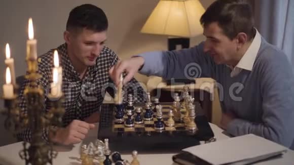 两个积极的白人男子在国际象棋比赛父亲和儿子晚上坐在桌子旁爱好生活方式视频的预览图