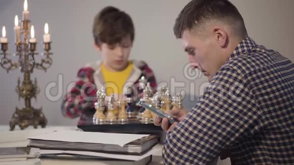 一位年轻的白种人使用智能手机的侧视肖像他的儿子坐在象棋的背景下心烦意乱视频的预览图