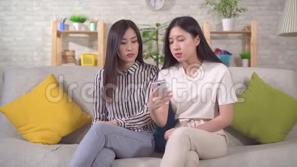 两位年轻的亚洲女性坐在沙发上看到了智能手机上令人震惊的内容视频的预览图