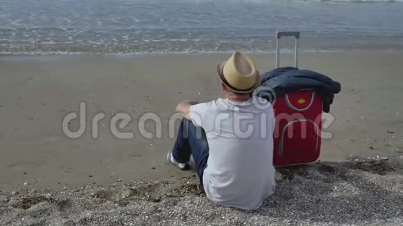 旅行者戴着帽子坐在海边的沙滩上旁边是一个红色的手提箱看着海浪视频的预览图