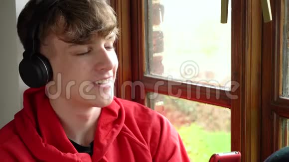 男青少年穿着红色帽衫坐在窗边听无线蓝牙耳机里的音乐视频的预览图