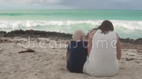 妈妈和儿子背对着镜头坐在海边的沙滩上视频的预览图