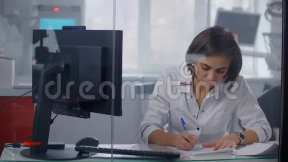 一位女医生坐在桌子旁用钢笔写处方给一个病人在玻璃墙后面视频的预览图