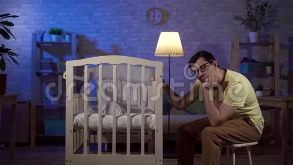 一个疲惫的年轻父亲睡着了一个新生儿晚上躺在婴儿床上视频的预览图