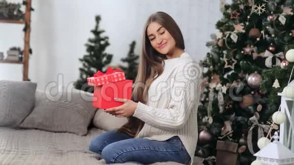 迷人的年轻女性微笑着拿着圣诞树附近的红色礼品盒红色相机宽镜头视频的预览图
