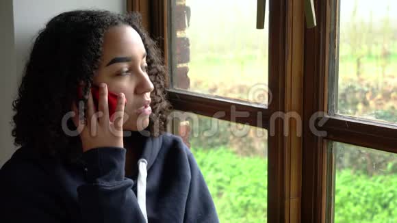 快乐的微笑美丽的混血儿非裔美国女孩少女坐在窗边用手机聊天视频的预览图