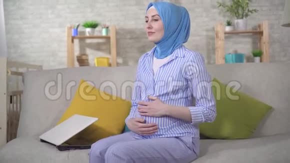 年轻的穆斯林孕妇戴着头巾坐在沙发上会感到头痛视频的预览图