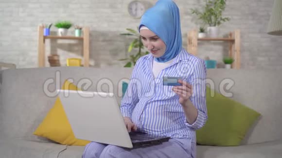 戴头巾的年轻穆斯林孕妇使用笔记本电脑和银行卡视频的预览图