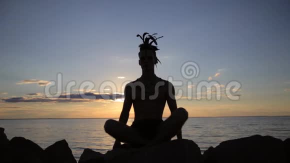 日落时分一个头上戴着美洲印第安人羽毛摩霍克配件的男子练习瑜伽的剪影视频的预览图