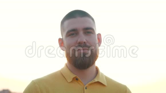英俊的欧洲年轻人留着胡须穿着黄色马球衫站在明亮的阳光下的田野上的肖像视频的预览图