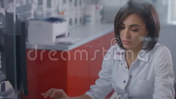 专业女医生穿着实验室外套在电脑上打字办公室里的玻璃墙后面坐着一个监视器视频的预览图