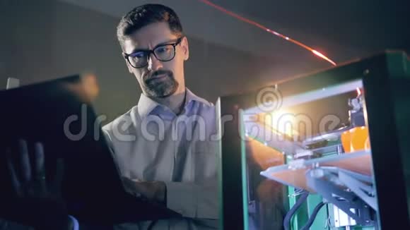 男工程师正在操作笔记本电脑同时观看3D打印机3D打印机用塑料打印物体视频的预览图