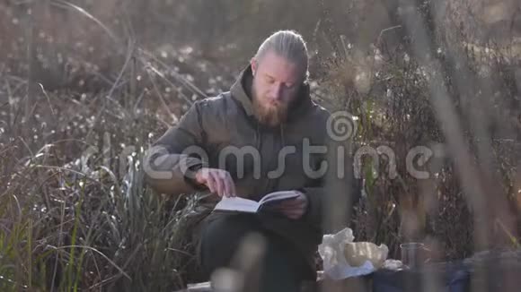 在秋天的森林里坐在树干上读书的有思想的高加索人的肖像男性游客度假视频的预览图