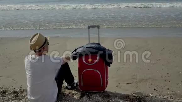 带着红色手提箱的游客坐在海滩上观看海浪视频的预览图