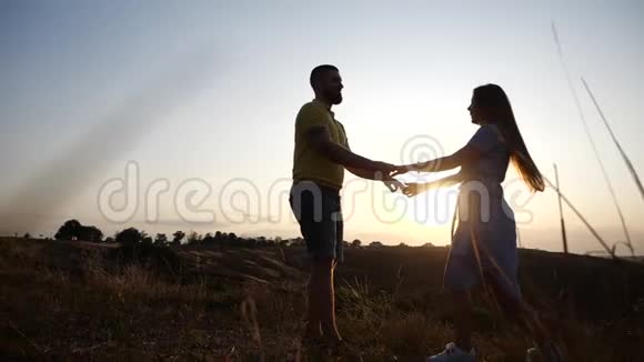 在黎明的背景下爱着年轻夫妇的女孩和男孩牵着手调情亲吻和拥抱在田野上慢慢地视频的预览图
