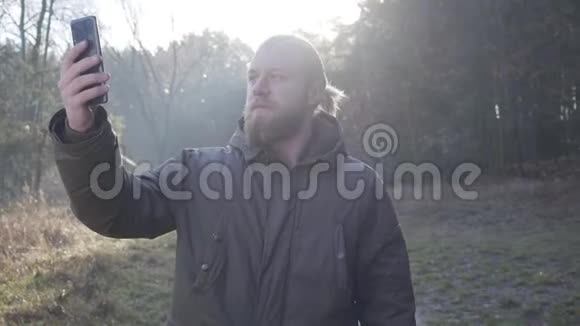 年轻的白种人留着胡子和马尾辫站在秋天的森林里打着视频电话成年男性游客视频的预览图