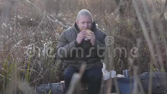 英俊的白种人坐在秋林的树干上吃热狗喝茶男性游客休息视频的预览图
